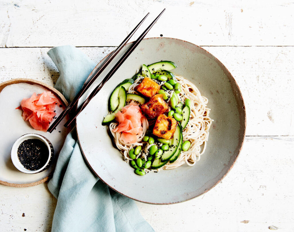 Overhead image of a bowl of soba noodles with tofu, ginger, and greens at a place setting on a white table.