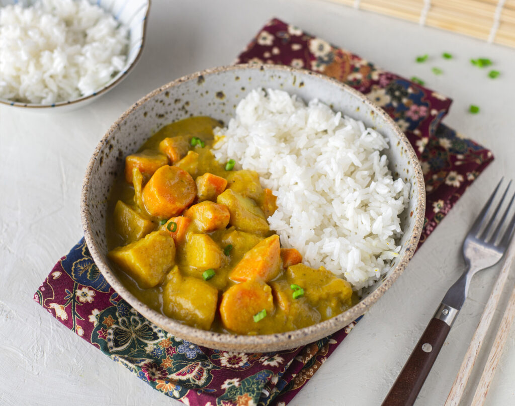 An overhead shot of a table setting with a bowl of curry and rice. The curry is in a golden sauce and shows big chunks of carrot and potato.