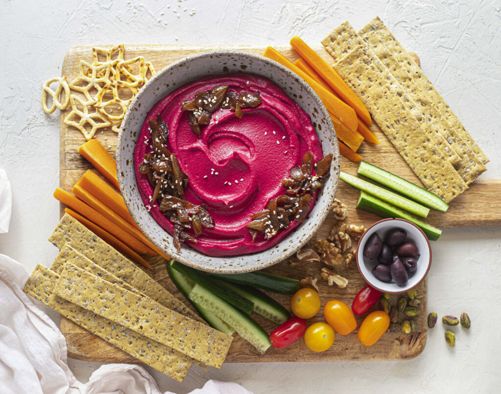 A vibrant purple beetroot dip sits in the centre of a platter, surrounded by crackers, raw veg, nuts and pretzels.