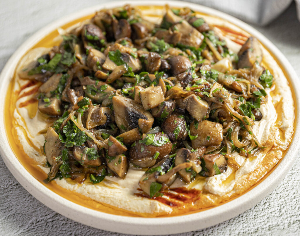 A close up of a bowl of creamy hummus topped with diced roast mushrooms and wilted greens.