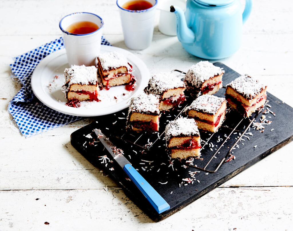 A shot of a table with a serving board of squares of jam-filled lamingtons, with two mugs of tea and a teapot in the background.