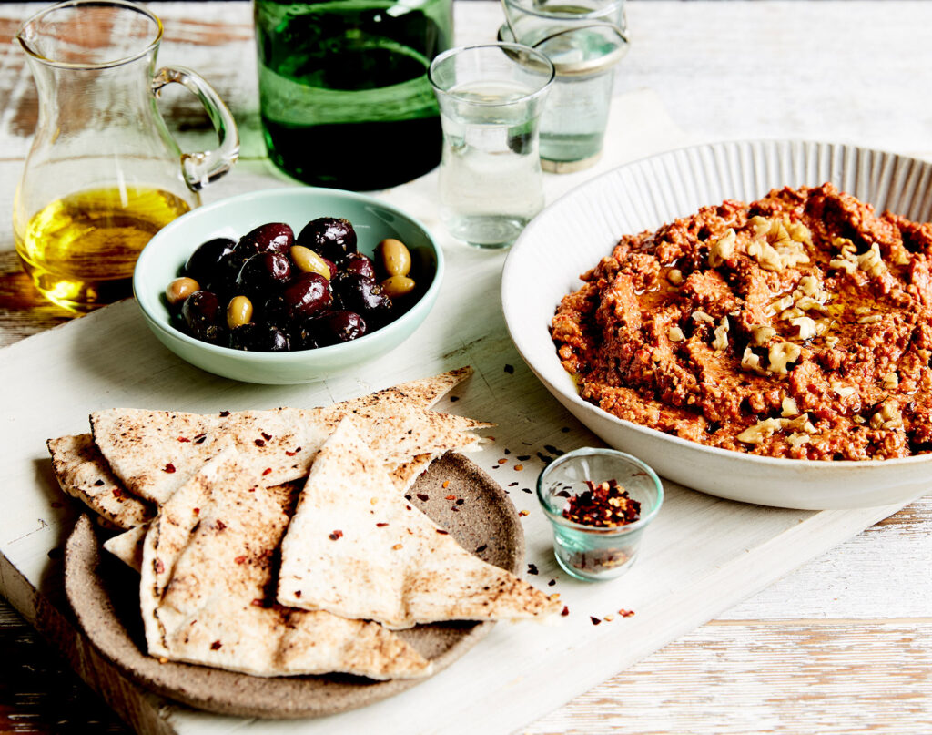 A chunky red dip sits to the left of a table setting, surrounded by pita bread, olives, chilli flakes, and a jar of olive oil.