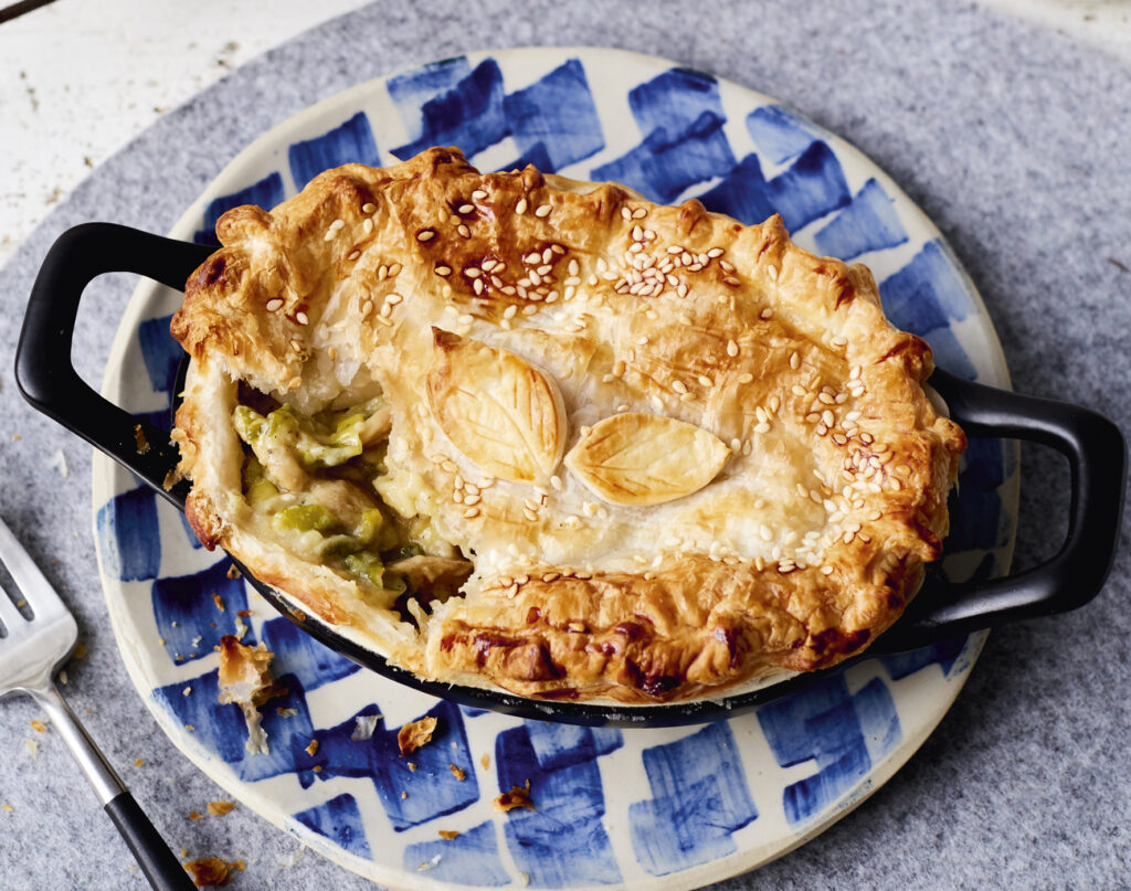 Overhead image of a beautifully decorated flaky pastry pie with leaf detailing. A portion of the pastry has been broken into with a fork to reveal a creamy leek and mushroom filling.