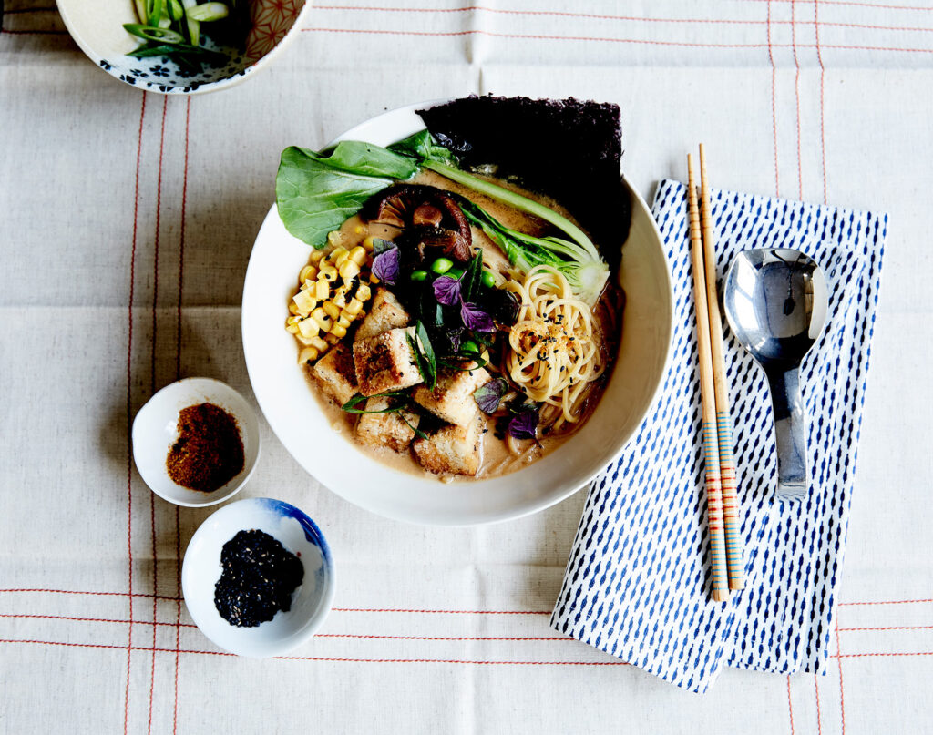 An overhead shot of a bowl of ramen noodles with tofu cubes at a table setting, with small bowls of seasonings to the side.