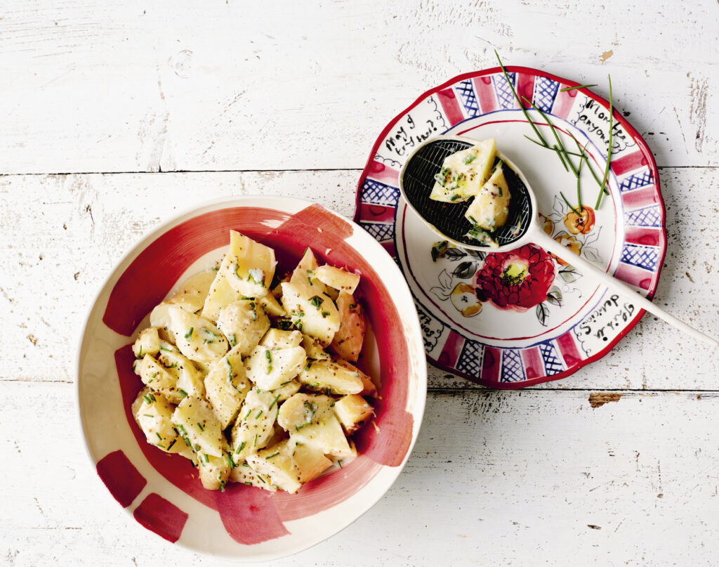 A serving bowl of creamy potato salad with a side plate sitting next to it. A salad server sits on the side plate with a small portion.