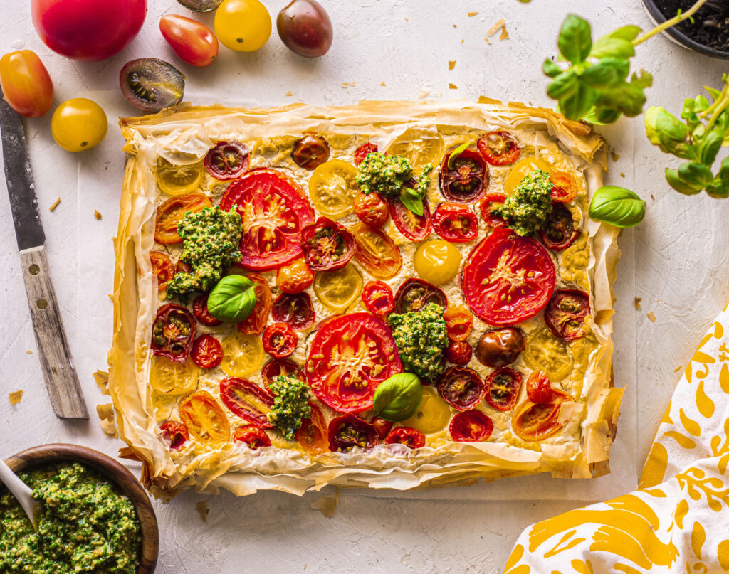 A rectangular tart made with flaky filo pastry topped with tomato, basil leaves and dollops of pesto.