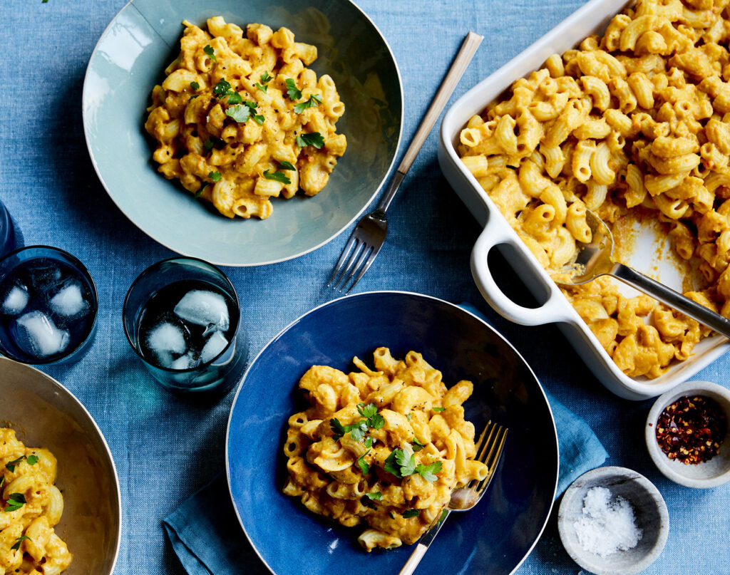 Overhead image of a serving dish or Mac & Cheese on a table, with several bowls served. There is a blue tablecloth underneath, and filled glasses sit to one side.