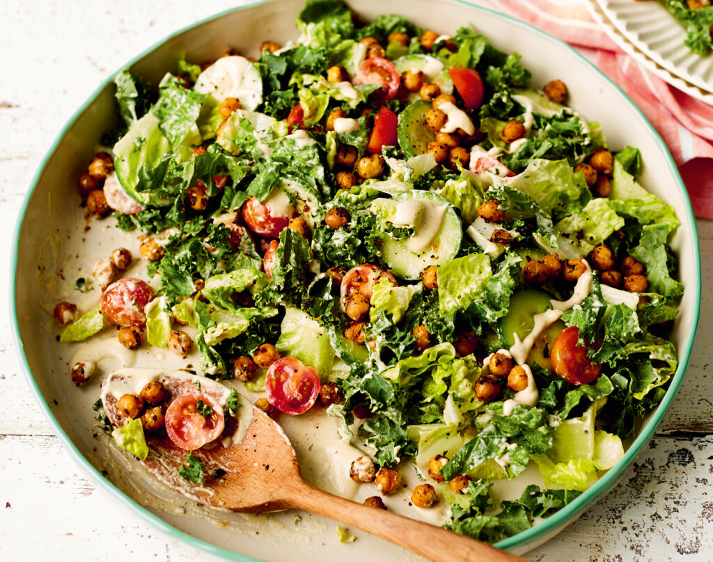 A big serving bowl of salad with a portion taken out. Plenty of greens with cherry tomatoes and roasted chickpeas are smothered in a creamy dressing.