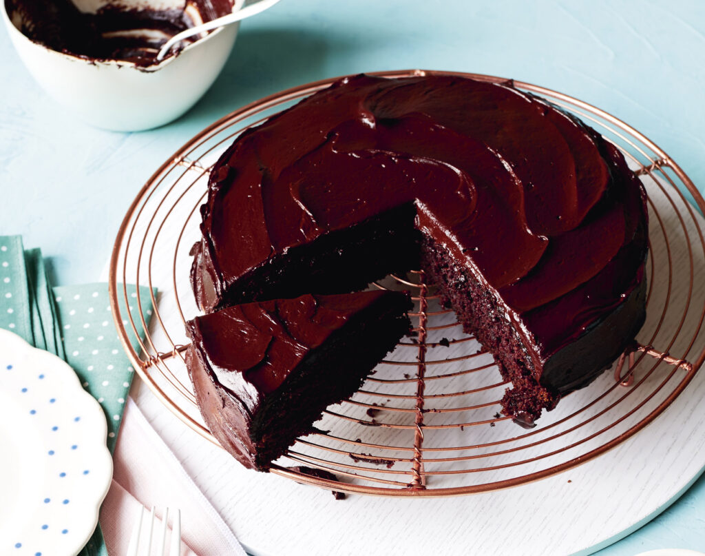 An overhead image of a rich chocolate cake covered in ganache on a cooling rack. A slice has been cut.