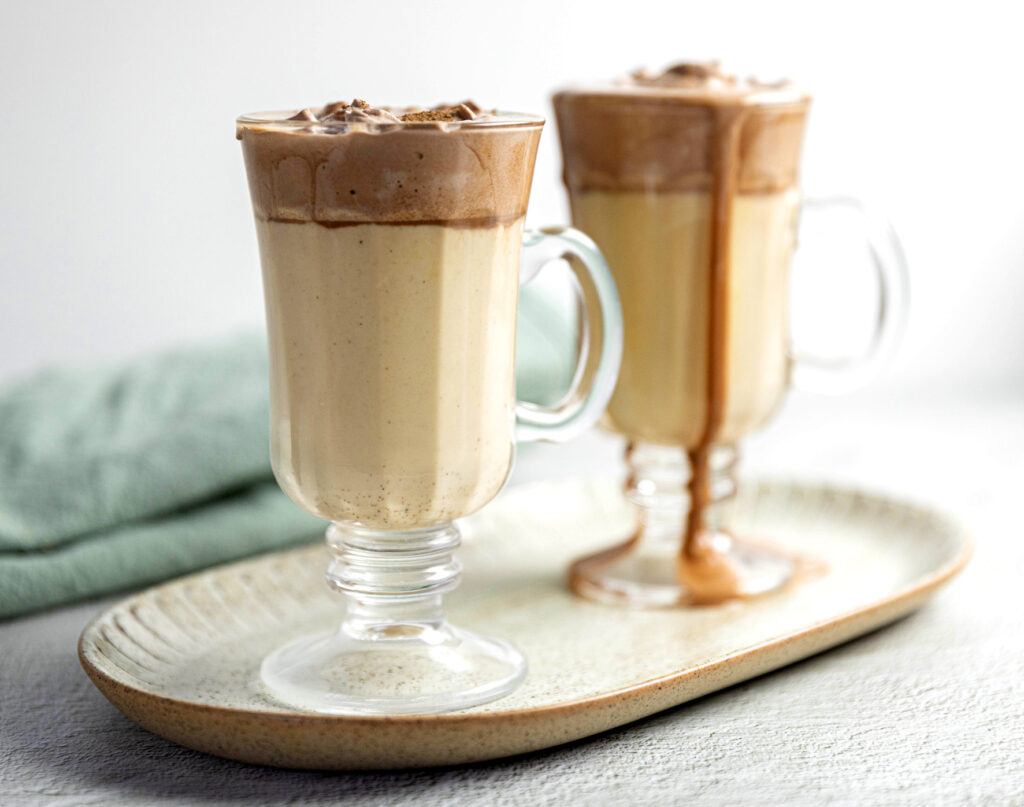 Two tall glasses with handles sit on a stoneware serving platter, full of oaty eggnog. The glass in the background is blurred with eggnog running down the side.