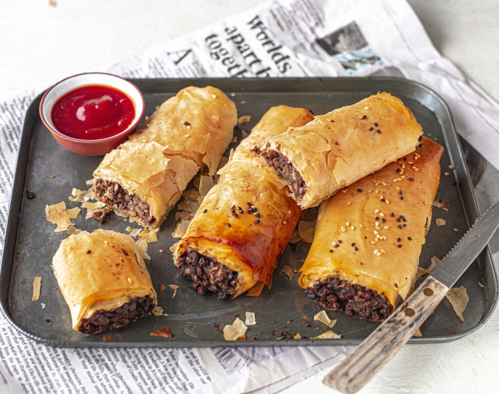 A rectangular tray of delicious looking flaky sausage rolls with a side of tomato sauce.