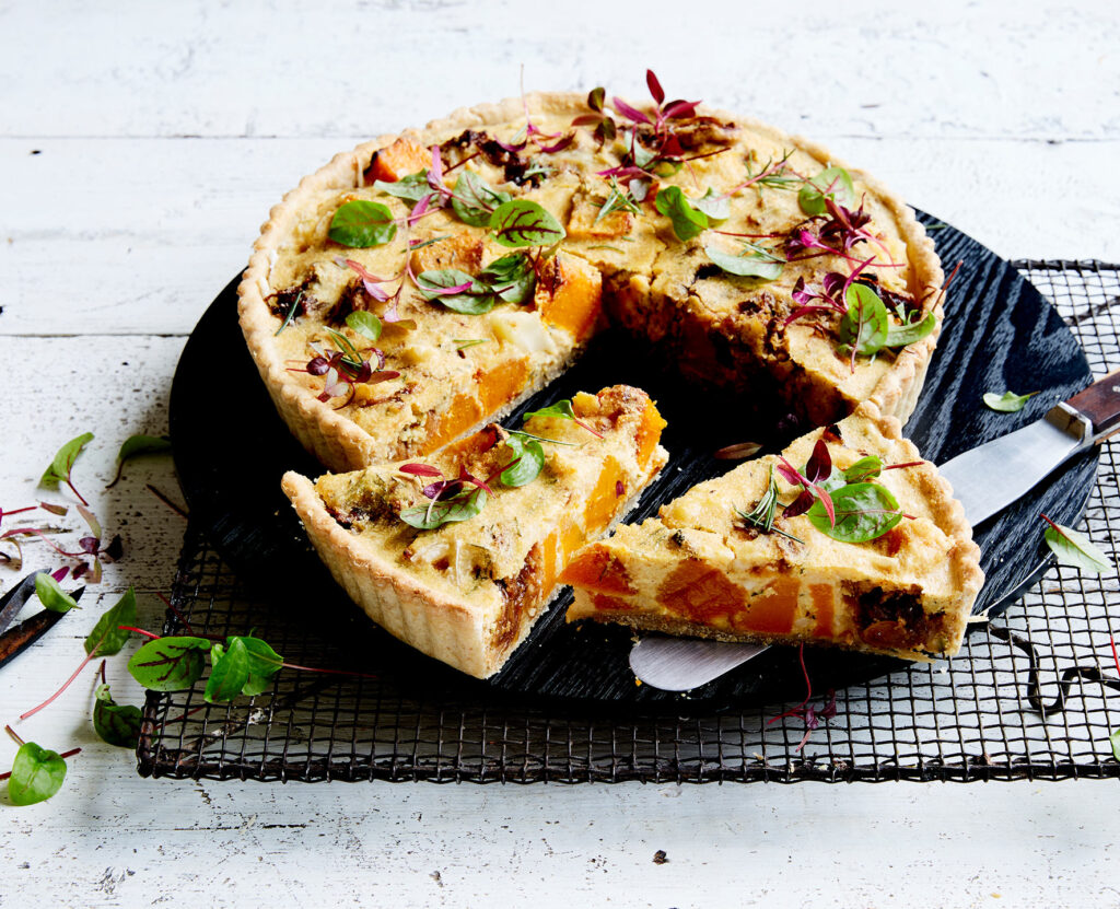 A round tart sits on a cooling rack, with two slices cut and sitting to the front of it, revealing the centre, which is not unlike a quiche.