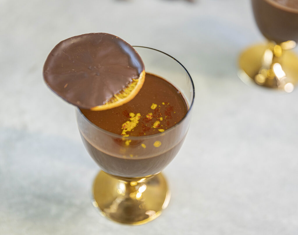 A goblet full of rich hot chocolate sits on a table. The image is taken overhead and the hot chocolate is garnished with orange zest. A chocolate covered slice of orange sits atop the glass.