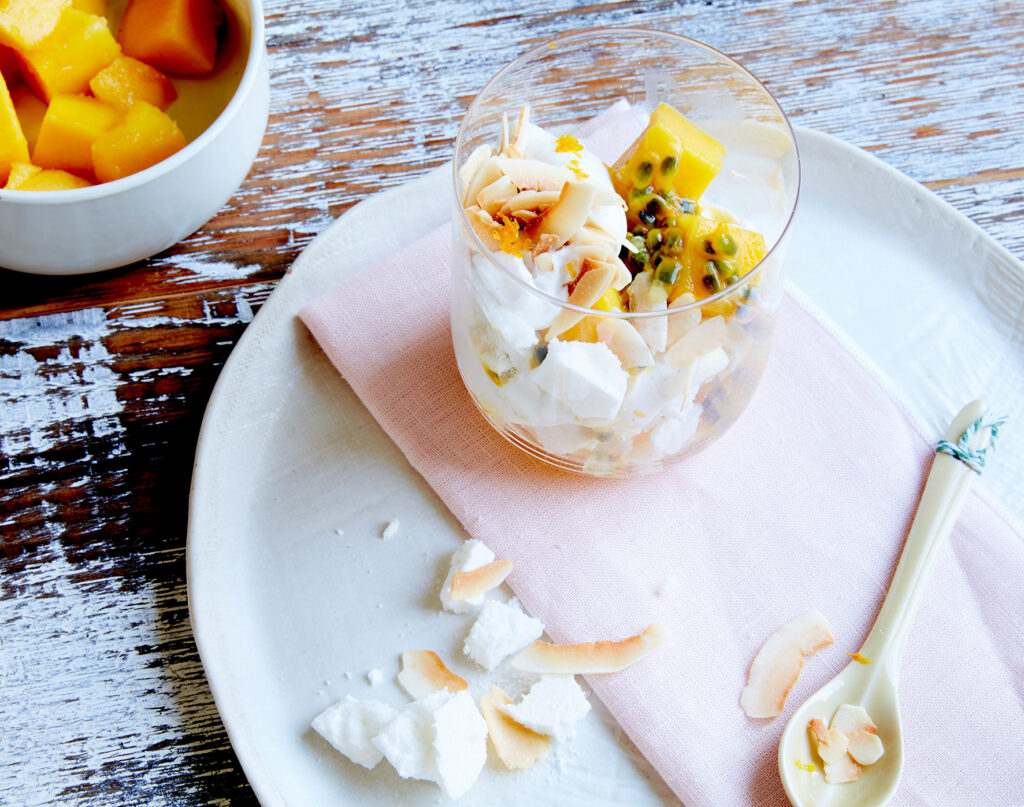 A clear glass filled with eton mess sits on a plate. It's topped with passionfruit, mango, and almonds.