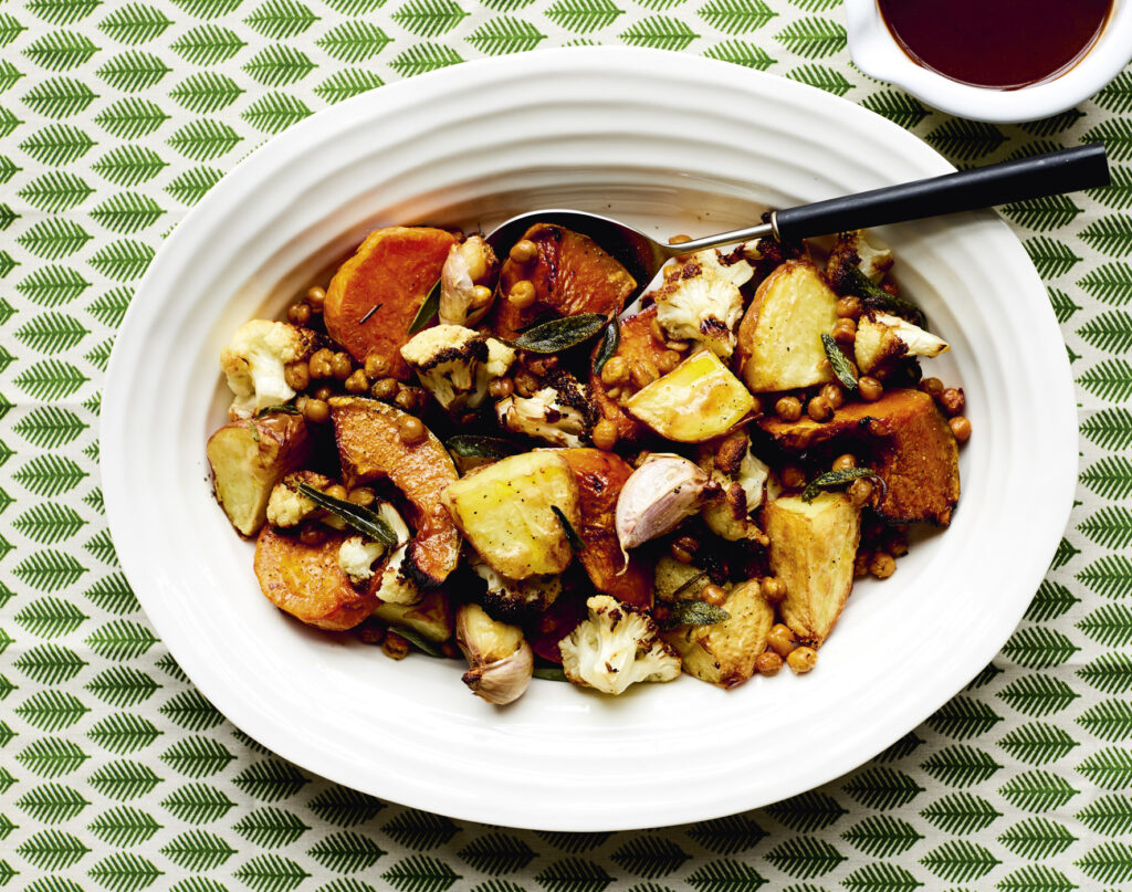 An overhead image of a bowl of herbed chickpeas sprinkled over roast potato and cauliflower. A 70s style patterned green and white tablecloth sits underneath.