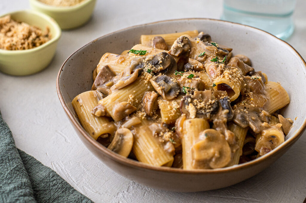 Close-up image of a bowl of a creamy mushroom rigatoni, garnished with herb crumb.