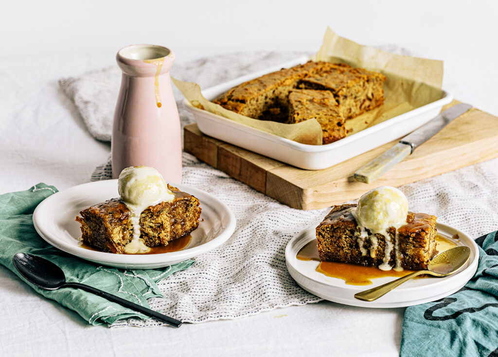 A slab of sticky date pudding on a serving board with two slices on plates in the foreground, topped with caramel sauce and ice cream.