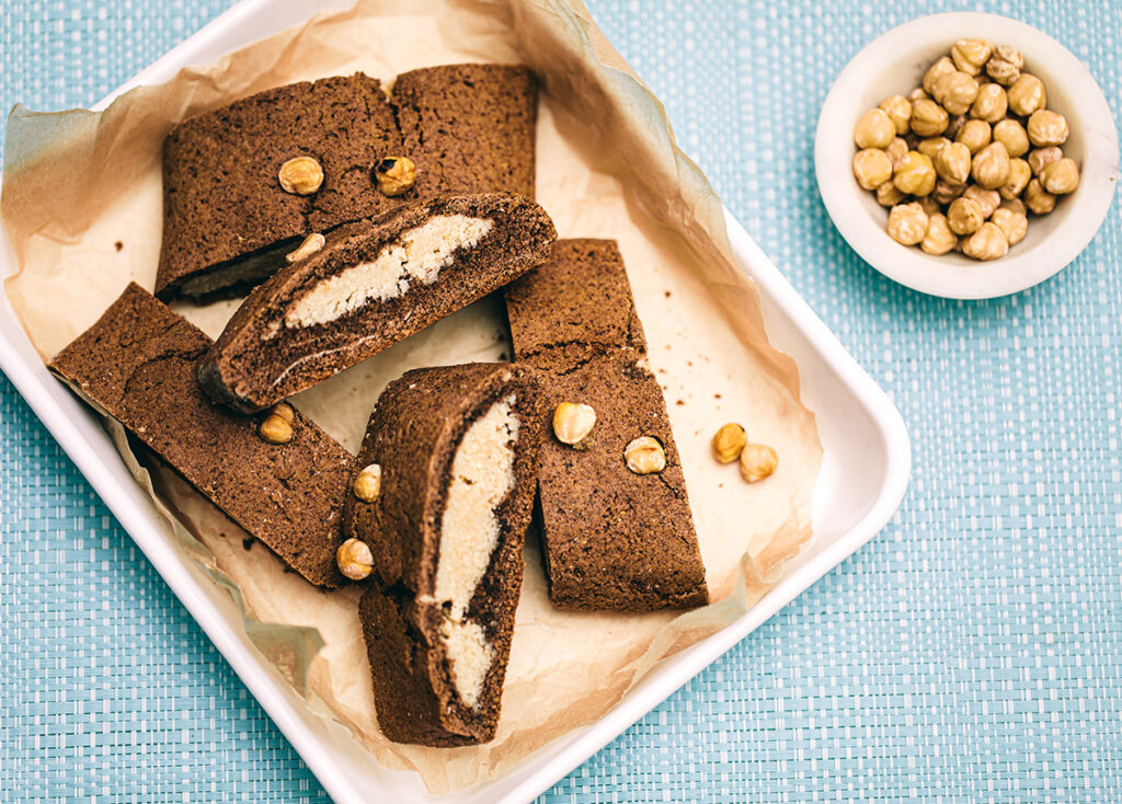 A rectangular tray with slices of Dutch spiced gingerbread log, revealing a creamy hazelnut filling in the centre. It is decorated with whole hazelnuts and a bowl or whole hazelnuts also sits to the side. 