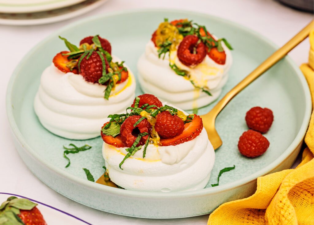 Three individual sized pavlova topped with berries and garnish sit on a green plate.