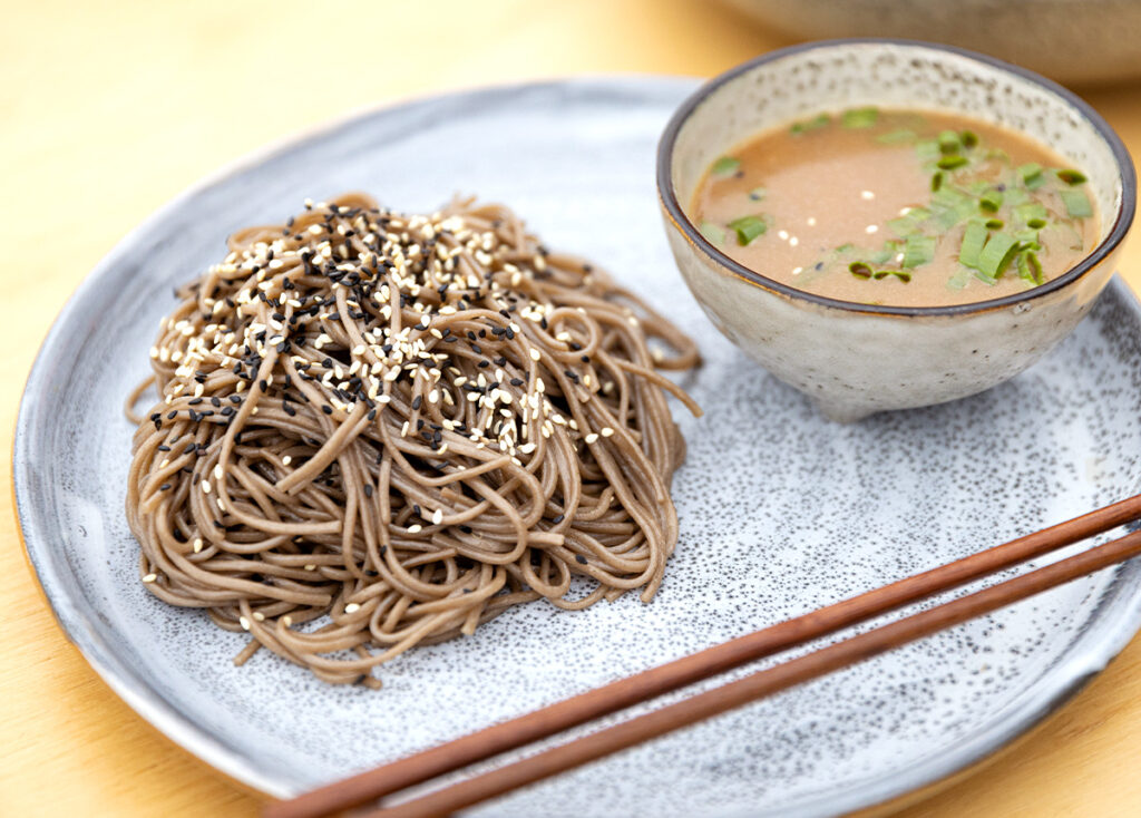 A neat pile of soba noodles sit on a plate with a round side bowl of dashi and chopsticks.