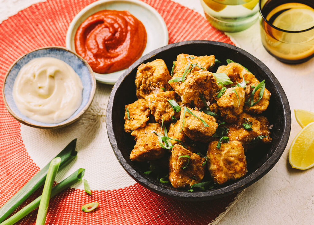 A bowl of perfectly crisp tofu bites at a table setting with several condiments to the side.