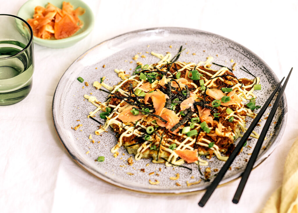 Image of a savoury Japanese pancake on a plate at a table setting, with a pair of chopsticks to the side.