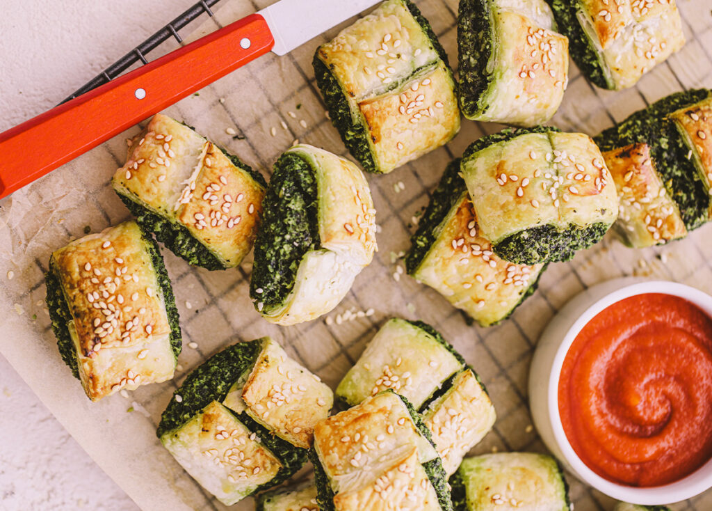 A tray of part-sized spinach and feta rolls with flaky pastry. A side of sauce sits to the side. 