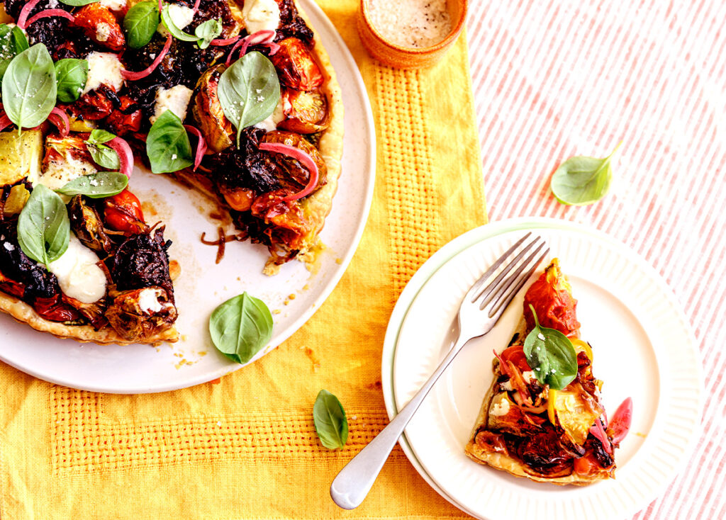 A round tart on a plate with a slice taken out, the slice sitting on a smaller plate next to it. The top is decorated colourfully with tomato and leaves of basil.