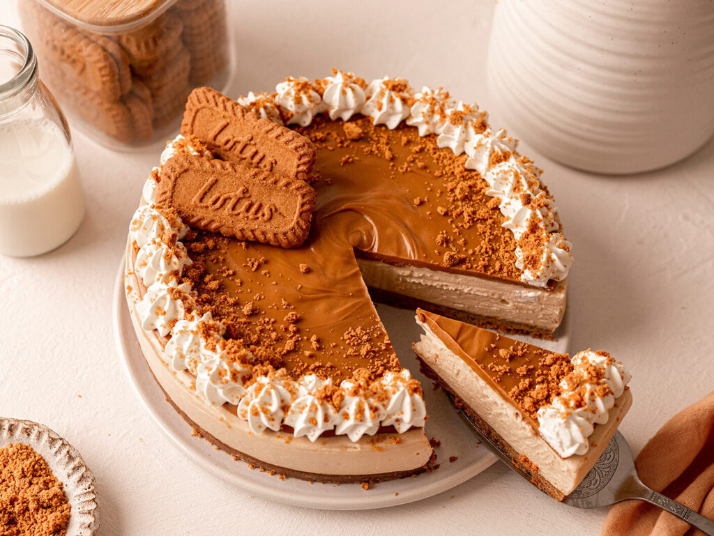 An overhead image of an iced Biscoff cheesecake. A slice is being pulled out on cake server with a hand in shot.