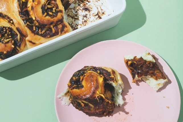 A freshly baked oven dish of Vegemite & Cheese Scrolls with one taken out on a plate beside it and pulled apart. On a green background.