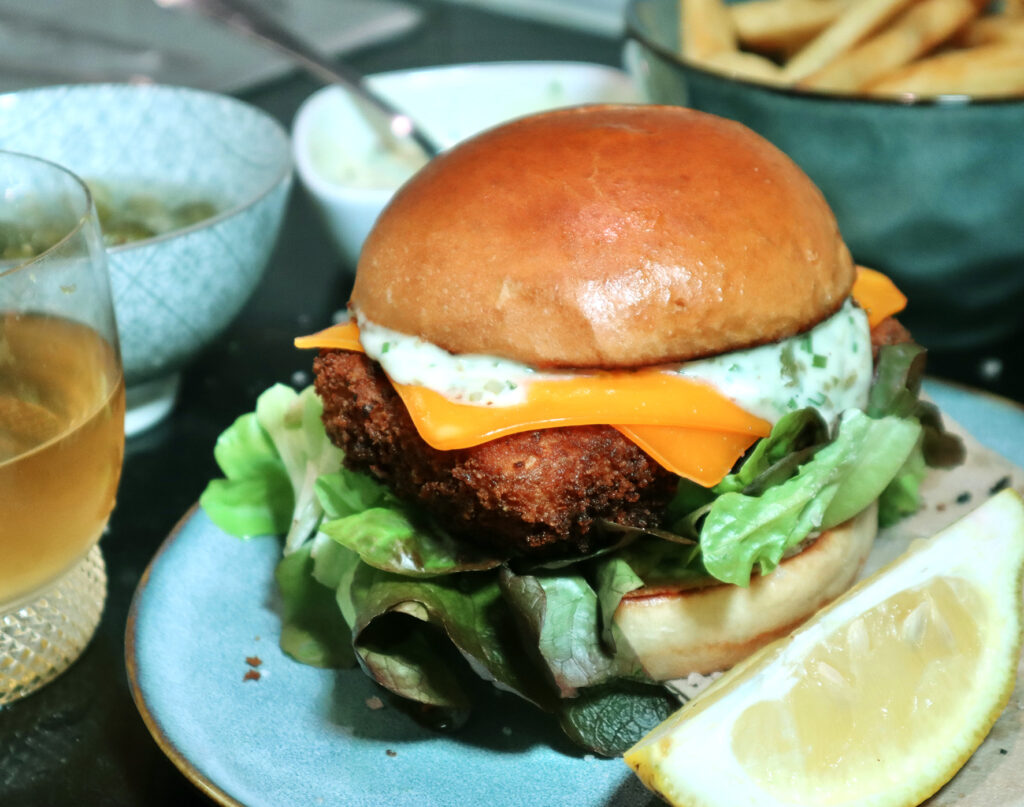 Delicious looking burger with crumbed vegan 'fish', cheese, tartare sauce, and lettuce, served with a wedge of lemon. 