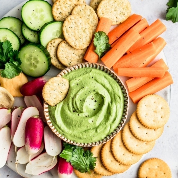 A creamy green dip sits at the centre of a platter, surrounded by crackers and raw veg. One cracker sits in the dip. 