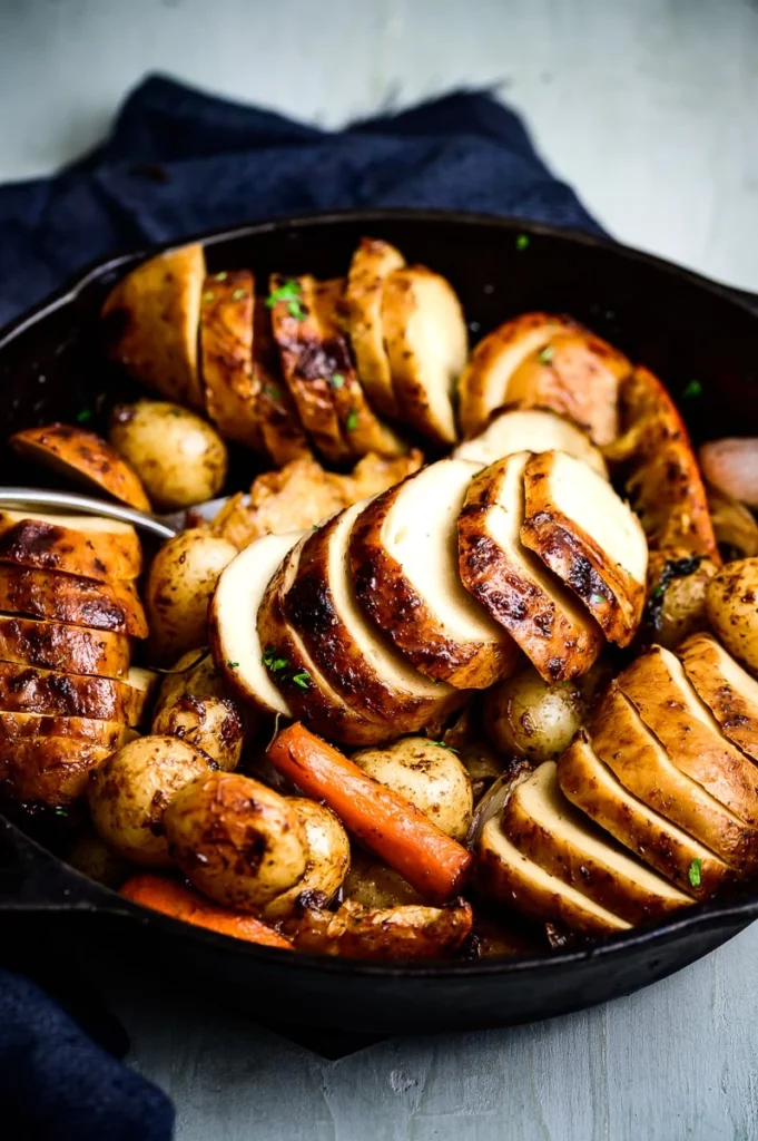 Image of thick slices of vegan roast chicken served in a black skillet with roast potato and carrot.