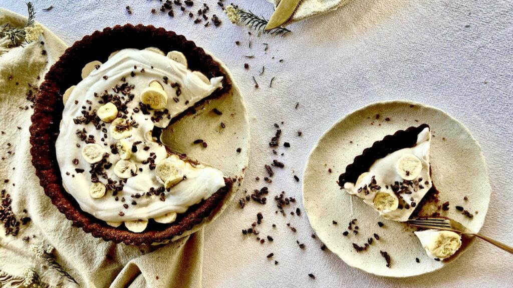 An overhead image of a cream-topped pie decorated with chocolate shavings and banana rounds with a slice on a plate to the right.