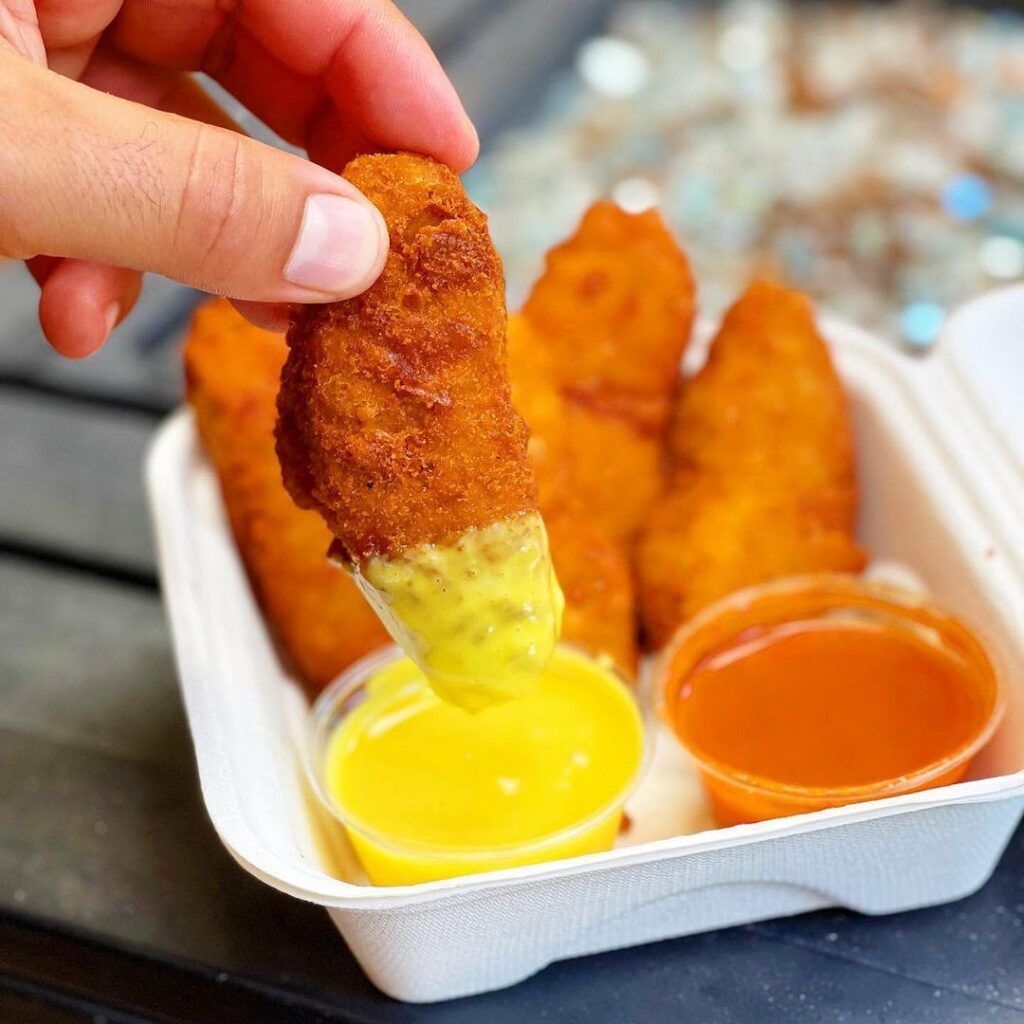A hand reaching over a takeaway container grabbing a vegan chicken tender and dipping it sauce.
