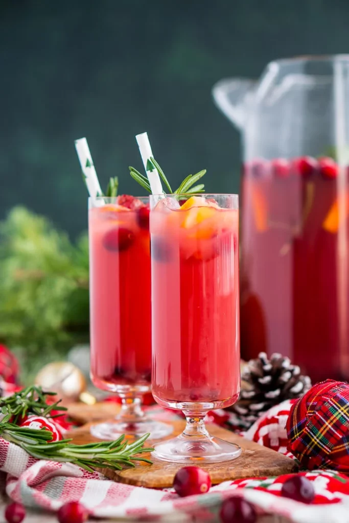 Two tall stemmed glasses full of red punch sit on a festively decorated table, with a large jug filled with punch in the background.