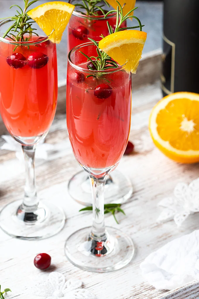 Three champagne flutes full of cranberry and orange mimosa sit atop a white tabletop, garnished with fresh cranberries and sliced oranges.