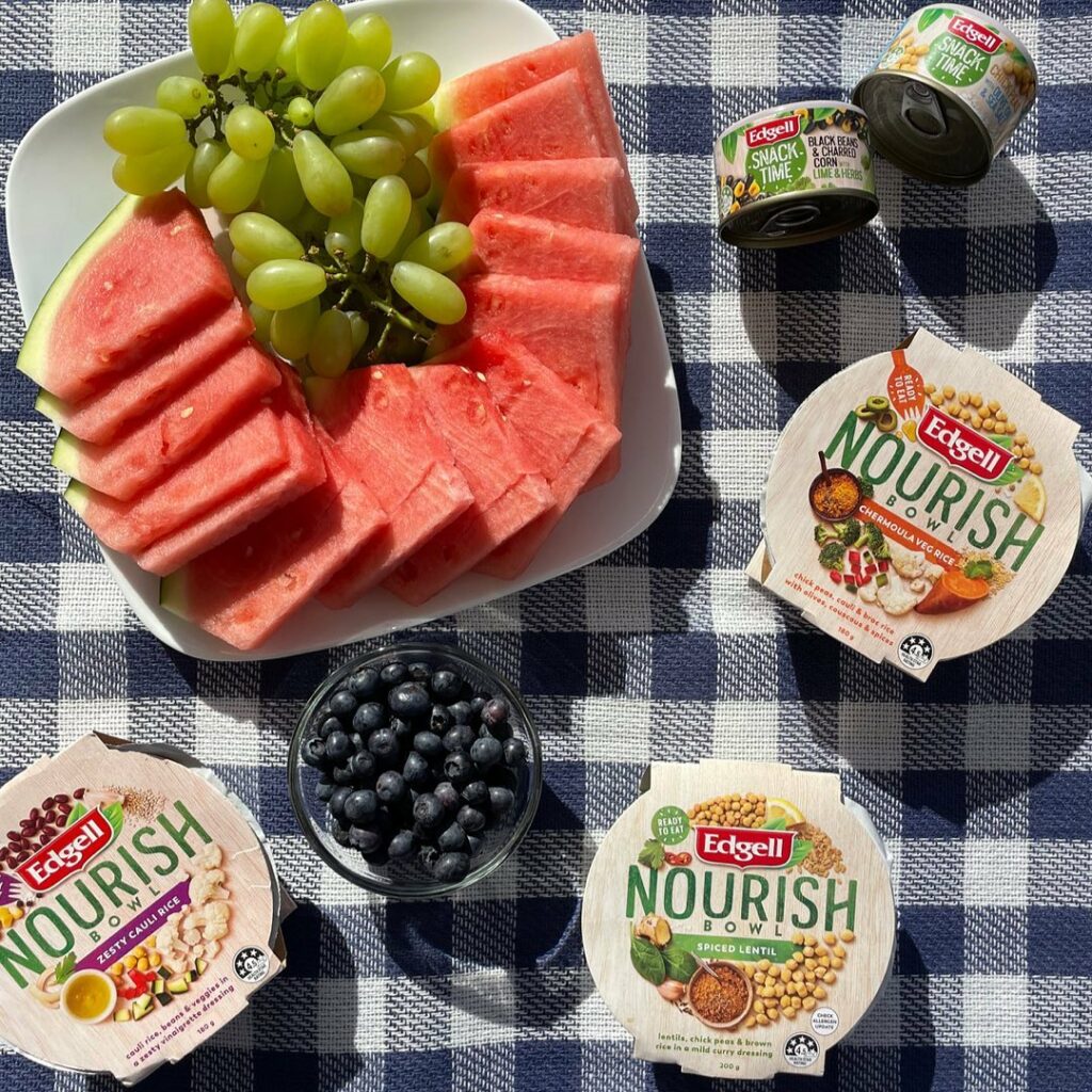 An overhead image of a picnic set-up, with a plate of watermelon and grapes, blueberries, and Edgell Nourish Bowls.