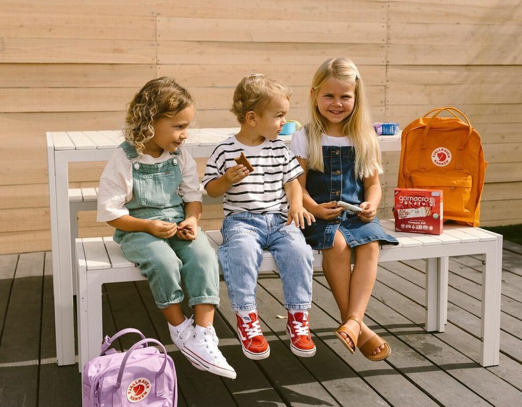 Three young children sit on an outdoor bench, snacking on Gomacro bars.