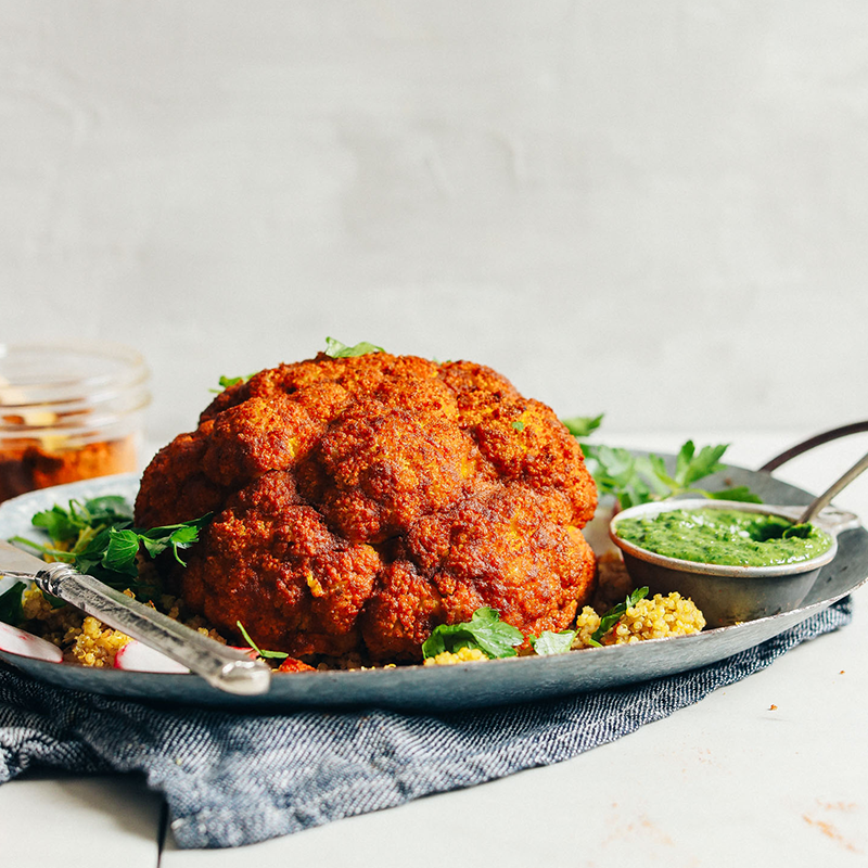 Image of a delicious looking roast cauliflower on a plate with some greens and a side of dipping sauce.