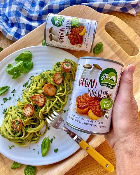 A hand holding up a tin of Nature's Charm scallops, with a plate of pasta on the bench below, topped with scallops.