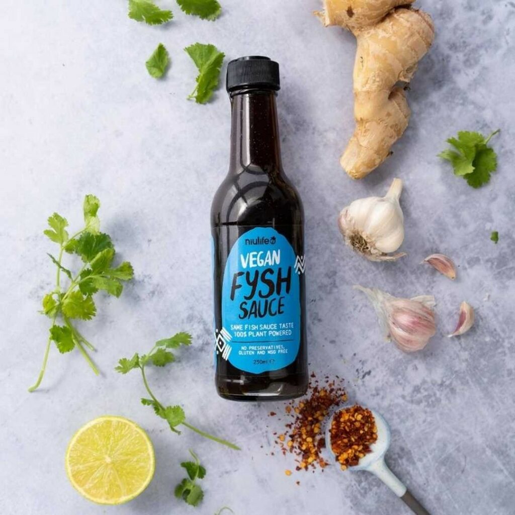 An overhead image of a bottle of fysh sauce, surrounded by ginger, garlic, lemon and spices on a benchtop.