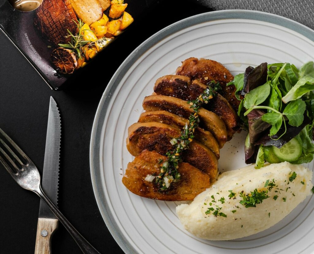 An overhead image of roast chicken sliced on a plate, with a side of mash and greens.