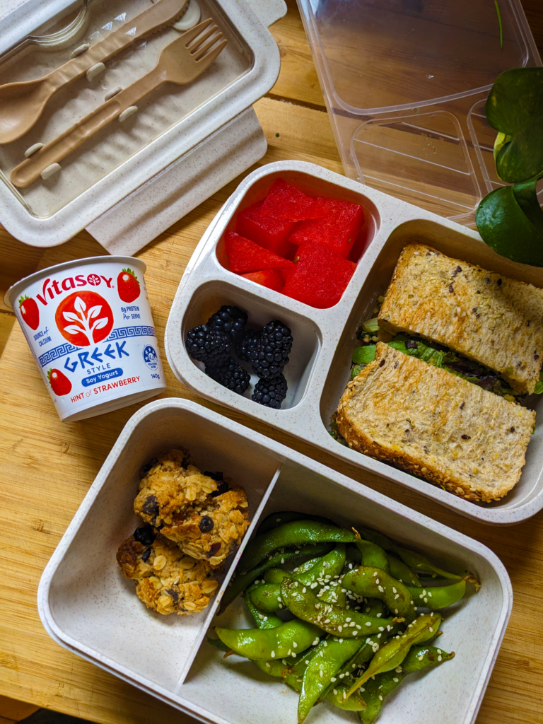 An overhead image of two bento boxes on a wooden bench filled with edamame, cookies, fruit, and a sandwich.