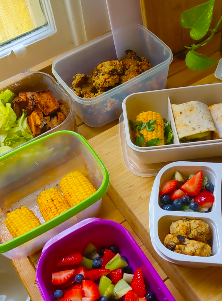 Overhead image of open tupperware containers on a bench filled with various pre-prepared plant-based lunchbox snacks. There are salads, chopped fruit, corn cobs, cookies, and wraps.