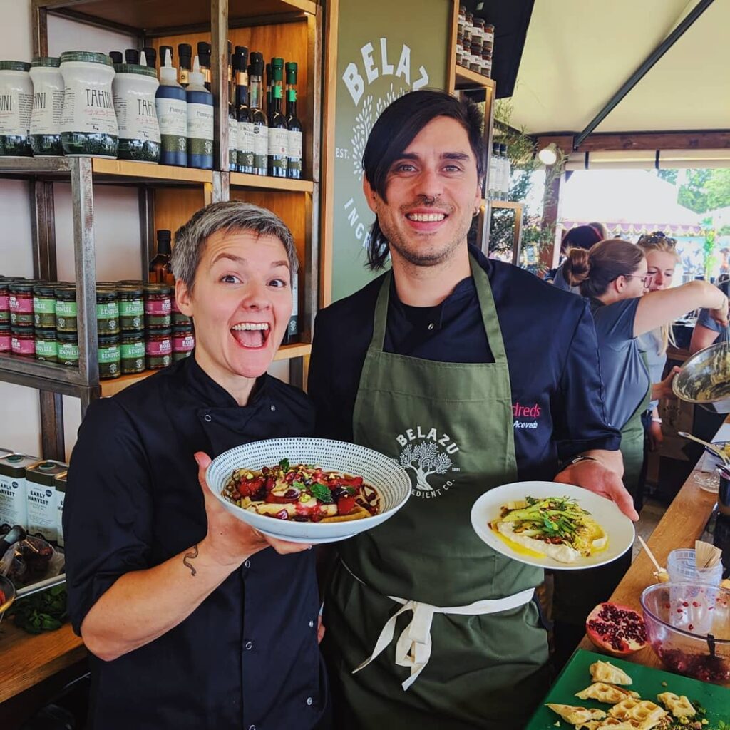 Two of the chefs at vegan restaurant, Mildred's, hold plates of food in front of them and smile at the camera.