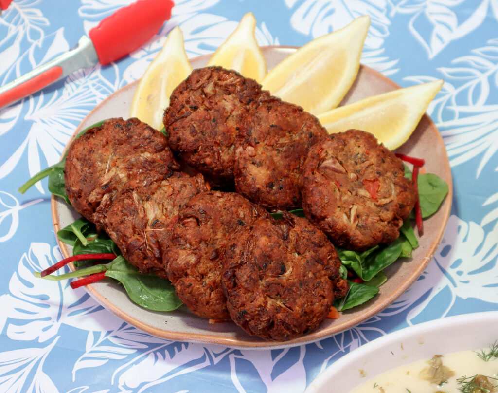 A serving plate of pan-fried vegan crab cakes on a bed of spinach served with lemon wedges.