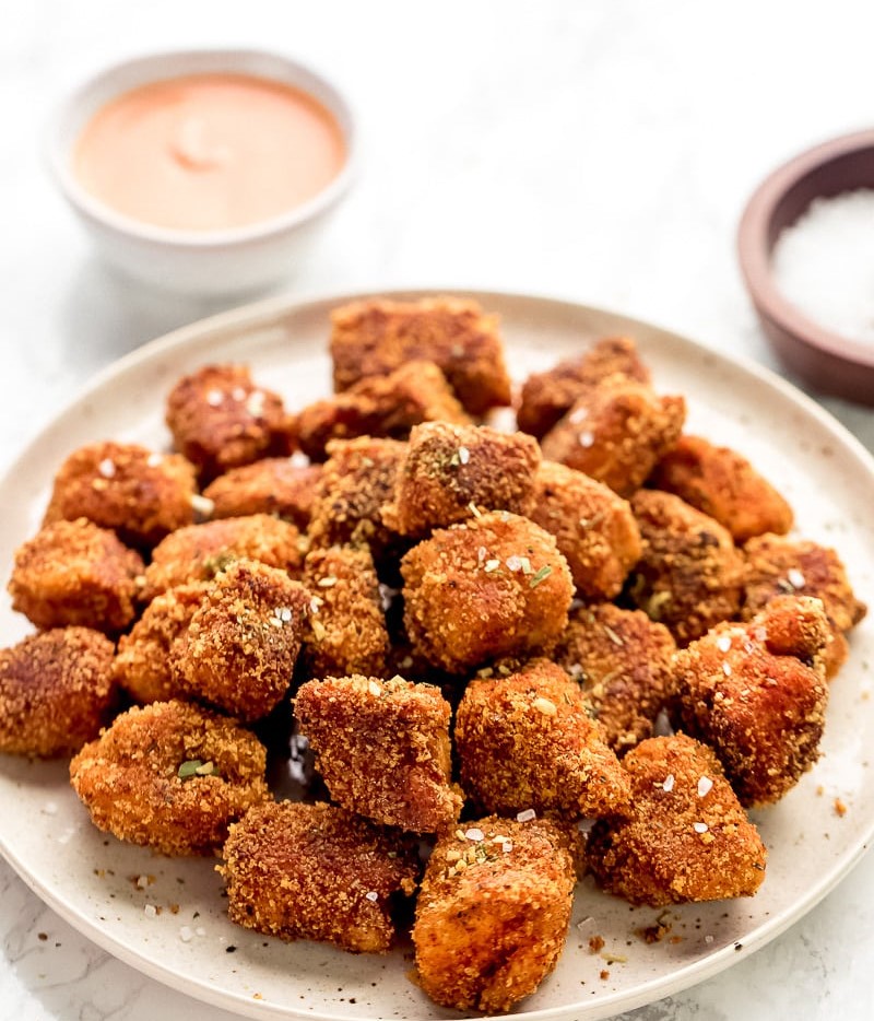 A close up of a plate of crispy tofu nuggets with a creamy dipping sauce visible in the background.