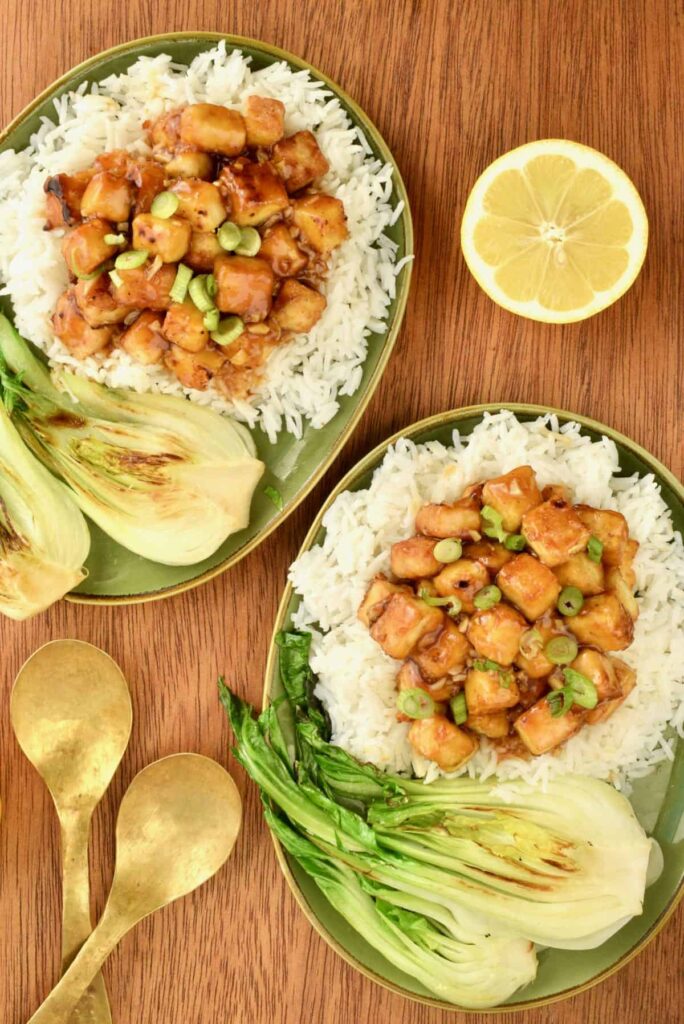 Overhead image of two plates of sticky lemon tofu nuggets served on a bed of white rice and a side of Asian greens.