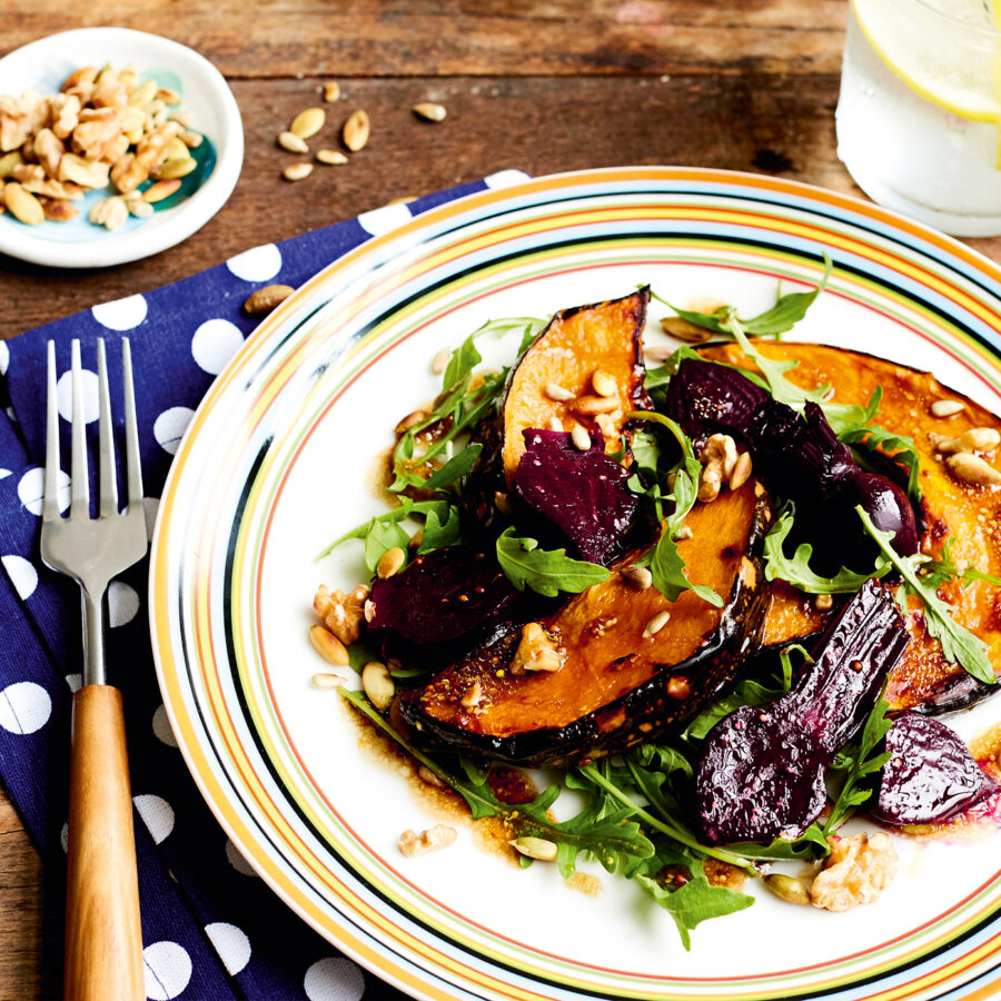 A plate of roasted pumpkin and beetroot salad with rocket, seeds, and nuts sits by a fork, serviette, and lemon water.